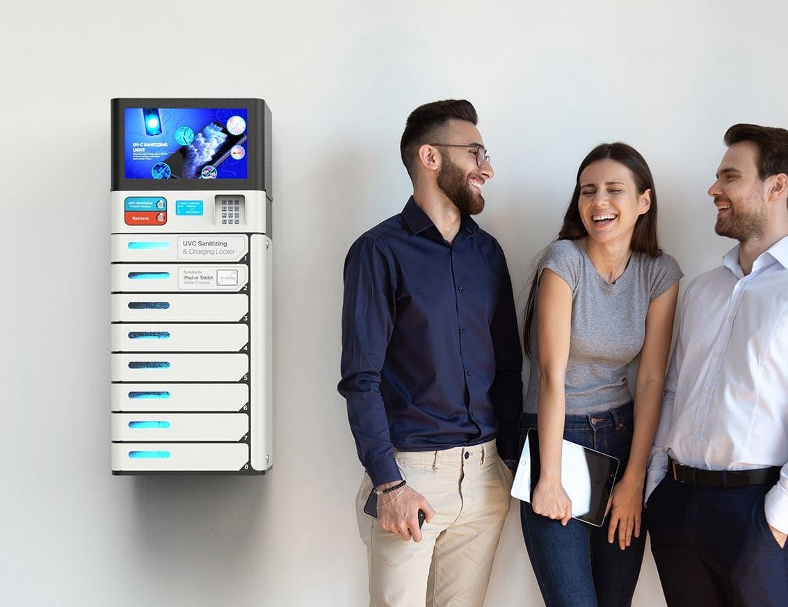 Three colleagues standing beside a wall-mounted charging locker with a display screen, in an office corridor