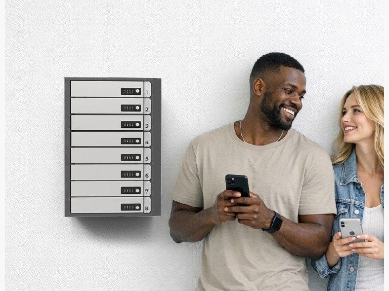 A compact phone charging locker mounted on a white wall in a public area, with two people standing beside it looking at a phone